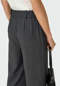 Femme portant un pantalon sur mesure gris foncé et un haut blanc, tenant un sac à main noir par sa bandoulière à côté d'elle.