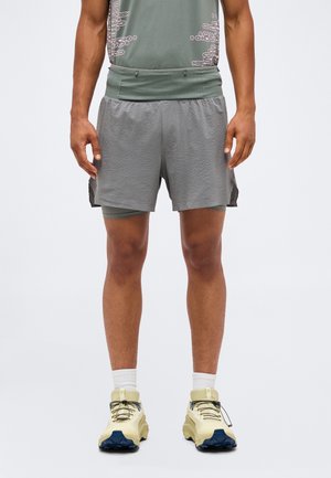 Homme portant un short de sport gris en couches, un t-shirt vert clair, des chaussettes blanches et des chaussures de course beiges, debout devant un fond uni.