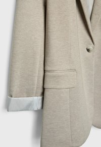 Béžový blazer s textúrovanou tkaninou; má jedno gombíkový zapínaním a pruhovaným detailom na manžetách. Strih prispôsobený postave a design s vykrojeným klopom.