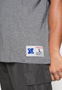 Camiseta de algodón gris con una etiqueta cosida con estrellas azules y blancas y la marca "Mitchell & Ness". Presenta un ajuste relajado y cuello redondo.