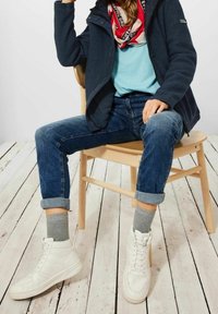 Dark blue fleece jacket, light blue t-shirt, rolled blue denim jeans, gray socks, and white high-top sneakers, seated on a wooden chair.