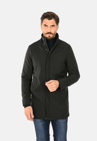 Cappotto impermeabile nero con colletto alto, bottoni a pressione e tasche laterali. Indossato sopra un maglione blu navy, abbinato a jeans azzurri.