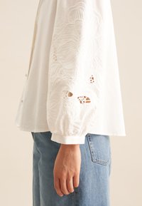 Blusa bianca con ricami floreali testurizzati sulle maniche, dotata di chiusura con bottoni e vestibilità comoda. Abbinata a jeans in denim azzurro chiaro.