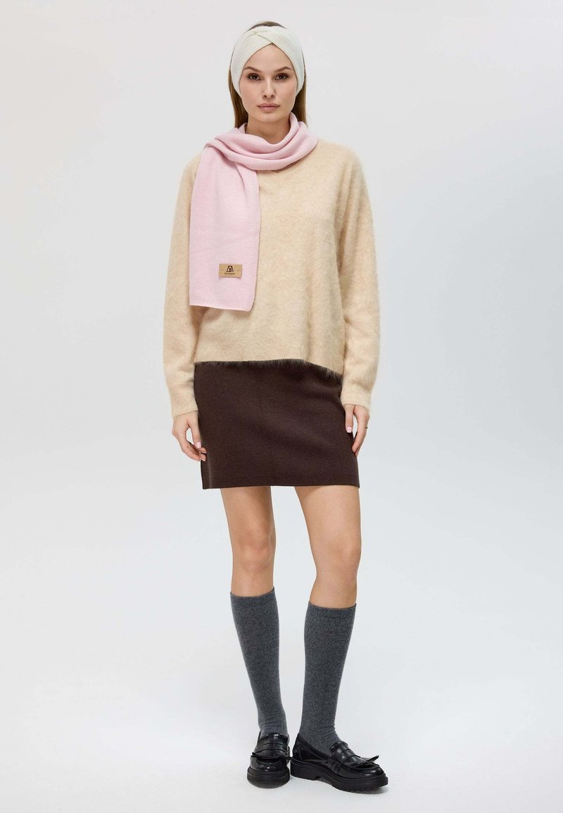 Pull en beige duveteux, jupe marron, écharpe rose clair, chaussettes grises jusqu'aux genoux et chaussures noires avec accents en nœud. Modèle se tient contre un fond clair.