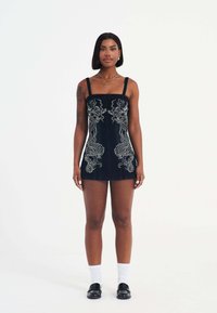 Zwarte denim mini-jurk met een strak ontwerp, met witte draak borduursels aan de voorkant en verstelbare bandjes. G gedragen met witte sokken en zwarte schoenen.