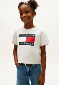 Szara bawełniana koszulka z czarnym i czerwonym prostokątnym logo z napisem "TOMMY" i "HILFIGER", zestawiona z niebieskimi dżinsami i czarnym paskiem.