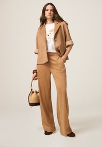 Veste en suede tan avec manches retroussées, associée à un pantalon assorti. T-shirt blanc avec motif graphique, sac à main marron à texture tissée, chaussures marron.