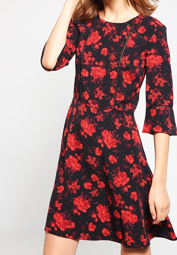 Femme portant une robe noire avec des motifs floraux rouges et des manches trois-quarts avec des poignets évasés, debout devant un fond uni.