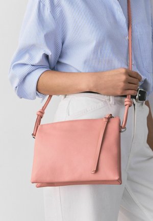 Persona, kas valkā zilu svītrainu krekli un baltas bikses, turot rokā mazu rozā ādas somiņu ar rāvējslēdzēju pāri ķermenim.