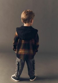 Laps, kes kannab musta kapuutsiga flanellsärki, millel on oranž ja kollane ruuduline muster, halli värvi cargo-pükse ja musti tossusid. Tagantvaade.