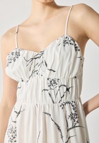 Robe blanche avec broderie florale noire, dotée de fines bretelles et de tissu froncé à la taille. Matière légère avec une texture douce.