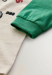 Licht beige shirt met een groen ribbelmanchet op de mouw. De stof lijkt zacht met een gladde textuur en bevat decoratieve elementen.