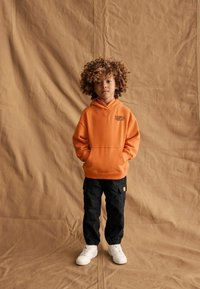 Felpa arancione con cappuccio, tasca frontale e logo, abbinata a pantaloni cargo neri e sneaker bianchi. Sfondo marrone texture.