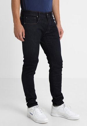 Mann trägt dunkelblaue, eng geschnittene Jeans und weiße Reebok-Sneaker, steht vor einem schlichten hellen Hintergrund.