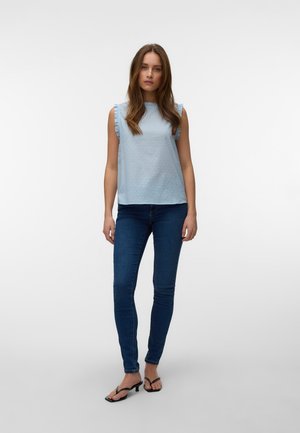 Femme aux longs cheveux bruns portant un haut sans manches bleu clair avec des volants, un jean skinny bleu foncé et des sandales à talons noires, debout.