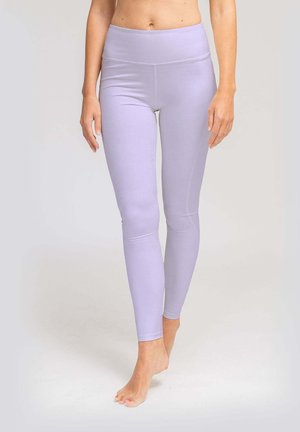 Frau trägt hoch taillierte lavendelfarbene Leggings und steht barfuß auf einem hellgrauen Hintergrund.