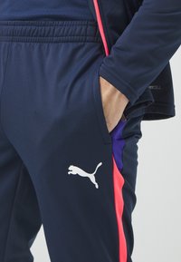 Mornarske športne hlače z rožnatimi in vijoličnimi stranskimi poudarki. Imajo belo Puma logotip, elastični pas in gladko, lahko tkanino.