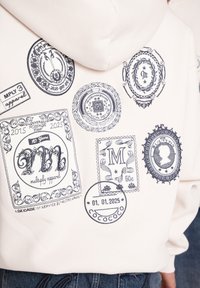 Hvid hættetrøje med forskellige sorte stempel designs, herunder tekst og dekorative elementer, på ryggen. Tekstelementerne fremhæver "Multiply Apparel" og datoer.