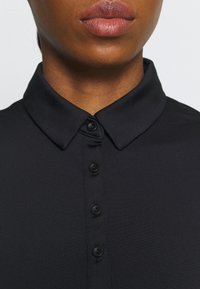 Czarna koszulka polo z zapięciem na guziki z przodu, o prążkowanej fakturze i klasycznym kołnierzyku. Posiada sześć czarnych guzików na listwie.