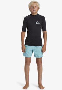 Tee-shirt de surf noir à manches courtes avec logo sur la poitrine, associé à un short de bain bleu clair. Debout pieds nus, avec les bras détendus le long du corps.