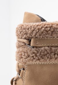 Brązowy zamszowy but z puszystym mankietem z owczej skóry, szczegółowym przeszyciem i metalowymi elementami. Mankiet jest teksturowany i pełny, oferując kontrast.