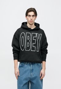 Fekete kapucnis pulóver, nagy fehér "OBEY" felirattal. Világoskék, bő szabású farmerrel párosítva, amelyen látható logó található a zsebnél.
