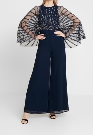 Kobieta w granatowym kombinezonie z półprzezroczystymi, błyszczącymi rękawami w paski i szerokimi spodniami, pozująca z rękami na biodrach.