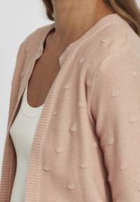 Cardigan rosa chiaro con una finitura testurizzata, caratterizzato da piccoli punti in rilievo e un colletto a costine, abbinato a una canotta bianca sottostante.