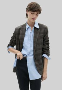 Blazer à carreaux marron sur une chemise à boutons bleu clair. Coupe slim avec des revers notch et des manches retroussées. Un pantalon noir complète la tenue.
