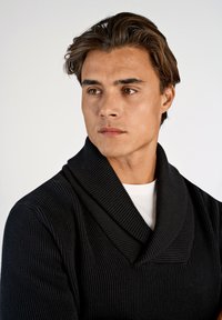 Zwarte geribbelde gebreide trui met een sjawlcol, laag over een wit shirt. Gladde textuur met subtiele details in het stofontwerp.