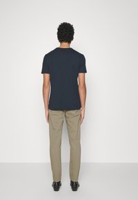 Man med lockigt hår står vänd mot en vit vägg iförd en svart kortärmad skjorta, beige byxor och svarta skor.