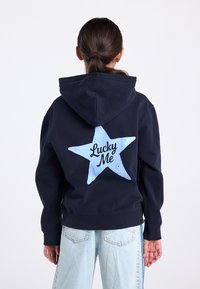 Marineblauwe hoodie met een grote lichtblauwe ster en "Lucky Me" gedrukt op de achterkant. Heeft een losse pasvorm en een kangoeroezak.