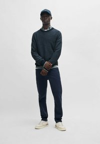 Dunkelblauer Strickpullover über einem hellgrauen Kragenhemd, kombiniert mit navyfarbener Hose und cremefarbenen Sneakers. Eine blaue Mütze rundet das Outfit ab.