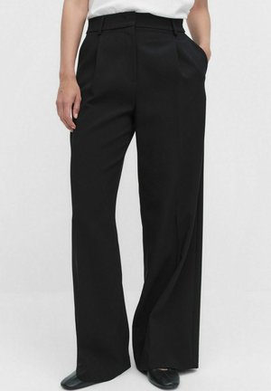 Zwarte wijde broek met een gladde textuur, plooien aan de voorkant en zakken, gecombineerd met een eenvoudig wit topje en zwarte schoenen.