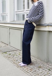 Pull rayé bleu marine et blanc avec une broche décorative en forme de fleur, associé à un pantalon en denim sombre à larges jambes et des mocassins brillants couleur lavande.