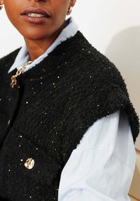 Veste noire texturée ornée de sequins dorés, avec un col rond et un bouton doré, portée sur une chemise bleu clair.