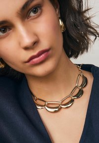 Frau mit kurzem dunklem Haar, die auffällige goldene Creolen und eine goldene Kettenhalskette mit großen, geschwungenen Gliedern über einem dunklen Oberteil trägt.