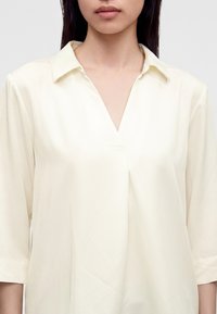 Femme portant un chemisier ample crème à manches longues avec un col en V, montré des épaules jusqu'au milieu de la poitrine.