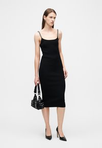 Γυναίκα που φοράει εφαρμοστό μαύρο midi φόρεμα με τιράντες από αλυσίδα, κρατάει μια μαύρη τσάντα με καρφιά και φοράει μαύρες μυτερές ψηλοτάκουνες γόβες.