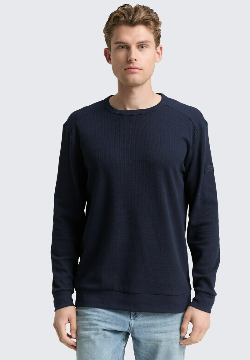 Marineblauwe rib gebreide sweatshirt met lange mouwen, ronde halslijn en een subtiele logo-detail op de linkerschouder. Casual pasvorm, zachte textuur.