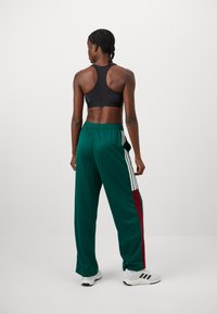 Zwarte sportbeha en groene trainingsbroek met witte en bordeauxrode zijstrepen. Soepele textuur, relaxed fit en elastische tailleband.