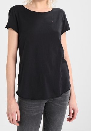 Vrouw draagt een effen zwart T-shirt met korte mouwen en vervaagde zwarte spijkerbroek, staand tegen een lichtgrijze achtergrond.