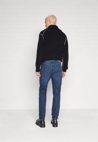 Schwarze Strickjacke mit hohem Kragen, versehen mit grauen Nähdetails, kombiniert mit blauen Jeans und schwarzen Schuhen. Rückansicht.