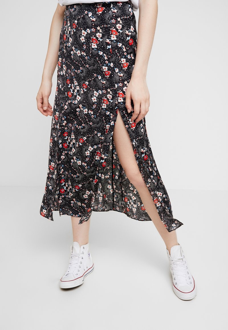 Zwarte bloemenrok met rode en witte bloempatronen, zijsplit, lichte stof, gedragen met witte sneakers. Middellange en vloeiende uitstraling.