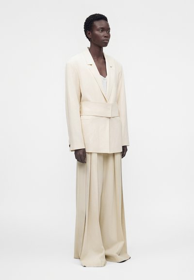 Άτομο που στέκεται, φορώντας ένα εκρού oversized σακάκι με φαρδιά ζώνη και αντιστοίχες φαρδιές παντελόνες, σε απλό λευκό φόντο.