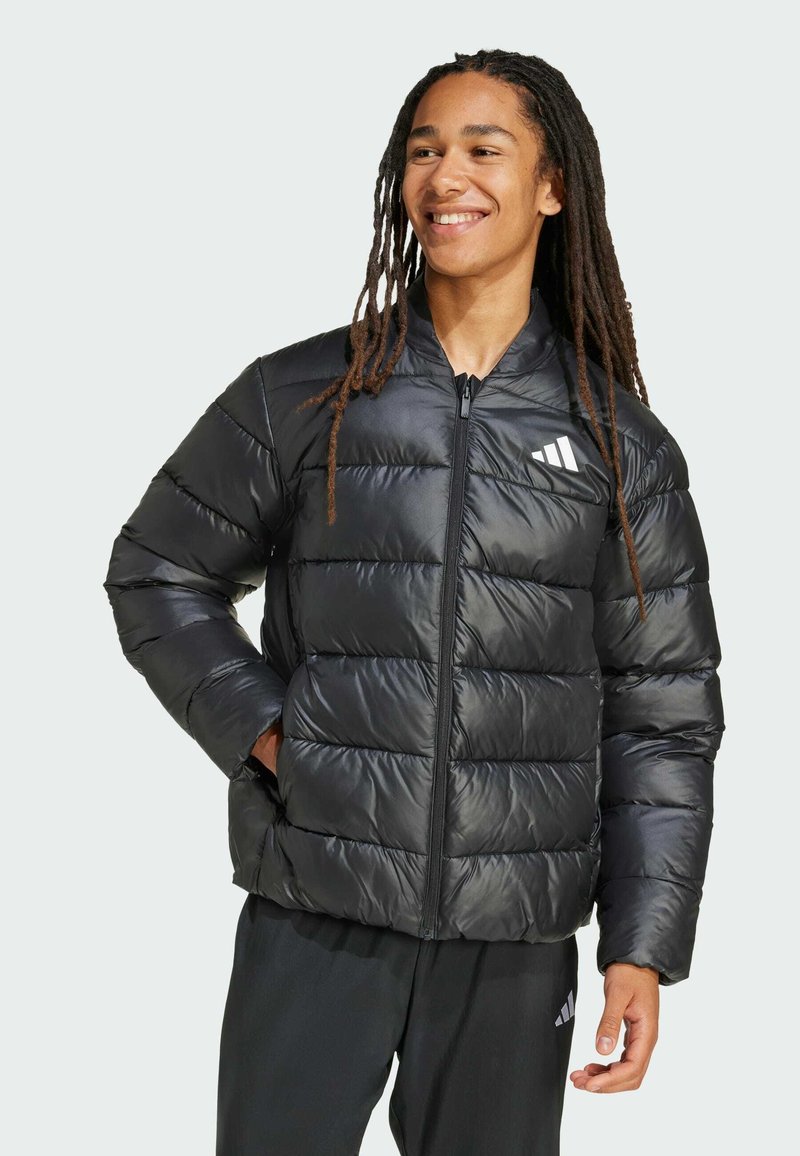 Nuori henkilö, jolla on pitkä tumma hius ja joka hymyilee, pukeutuneena mustaan paksuun takkiin ja tummaan housuun, seisoo vaaleaa taustaa vasten.