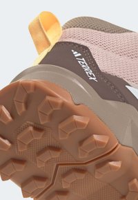 Turistická obuv Terrex s hnědým síťovaným svrškem, světlými růžovými akcenty, texturovanou gumovou podrážkou a charakteristickými výstupky pro trakci. Součástí je žlutý úchyt.