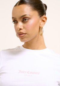 Balta medvilninė marškinė su mažuoju rožiniu "Juicy Couture SPORT" logotipu. Dideli auksiniai auskarai ir švelni, šviesi oda. Neutralus fonas.