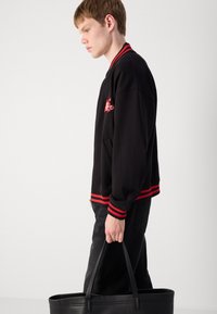 Black bomber jacket with red striped cuffs and collar; features an embroidered design on the chest. Model holds a black tote bag.