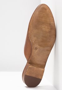 Semelle en cuir marron d'une chaussure habillée pour homme avec une semelle antidérapante texturée et la taille 37 indiquée, présentée en angle contre un mur d'angle blanc.